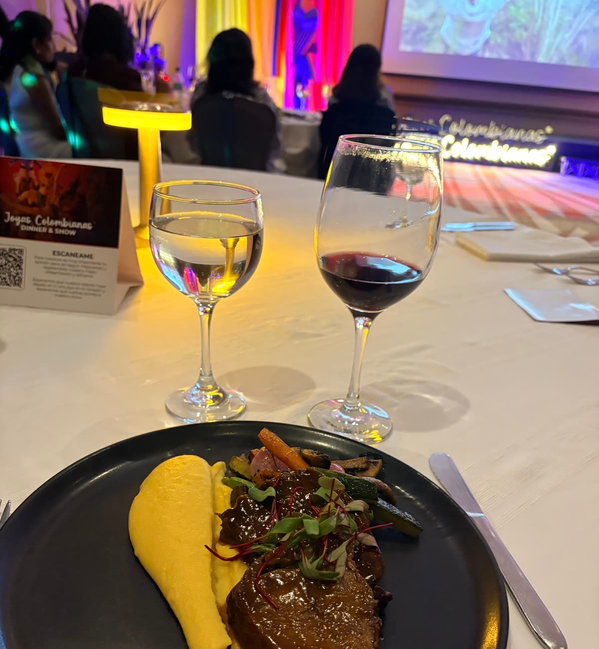 Mesa de Joyas Colombianas con un plato servido, copas de vino y agua, y el escenario del show al fondo