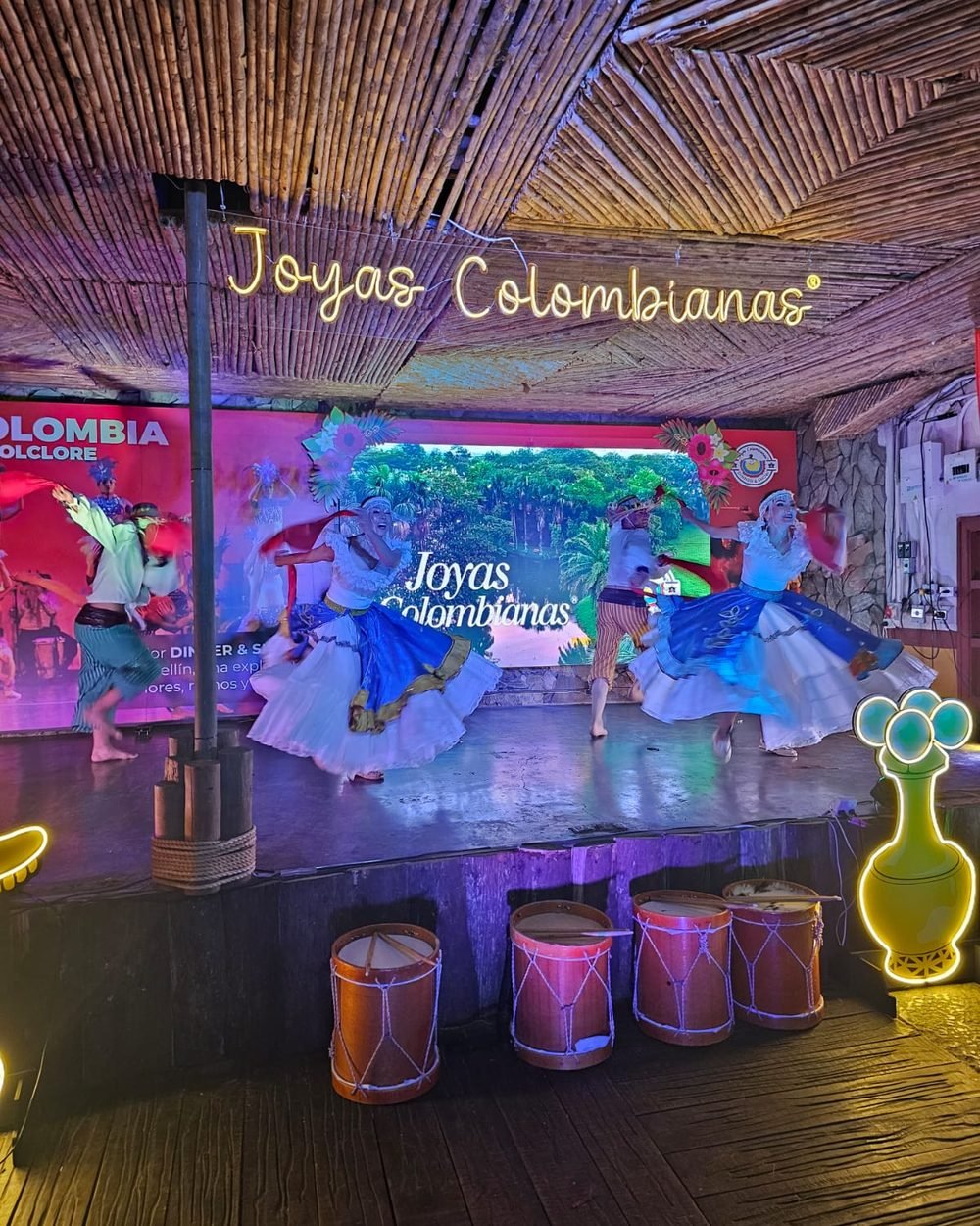 Bailarinas de Joyas Colombianas con faldas blancas y azules en el escenario, con tambores al frente