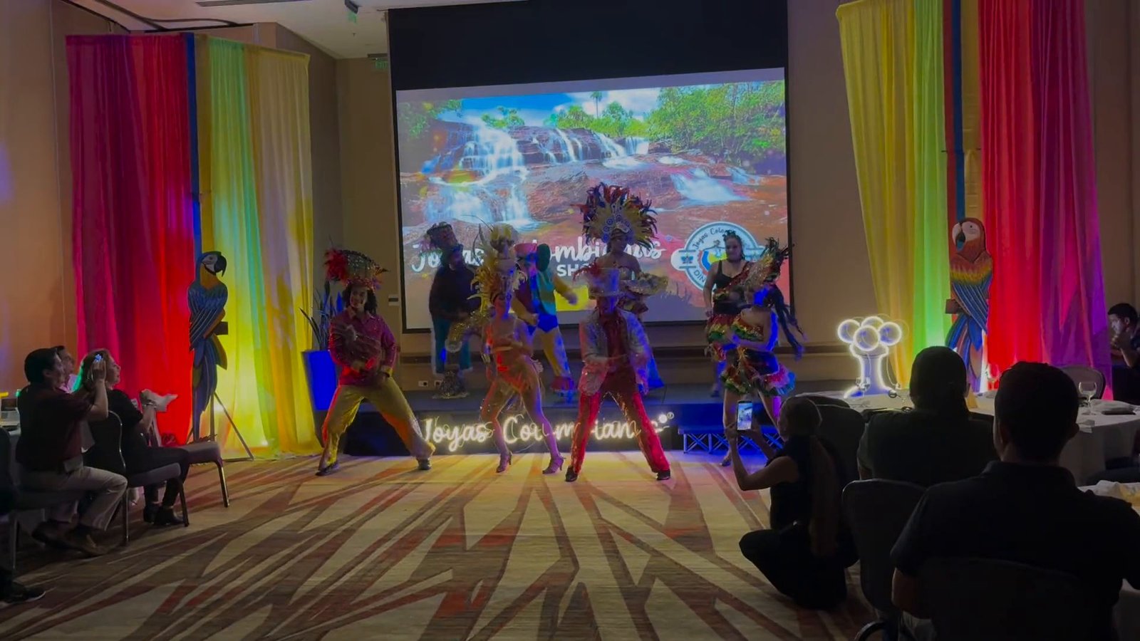 Bailarines de Joyas Colombianas con trajes de carnaval y tocados de plumas en el escenario, frente a los invitados en sus mesas